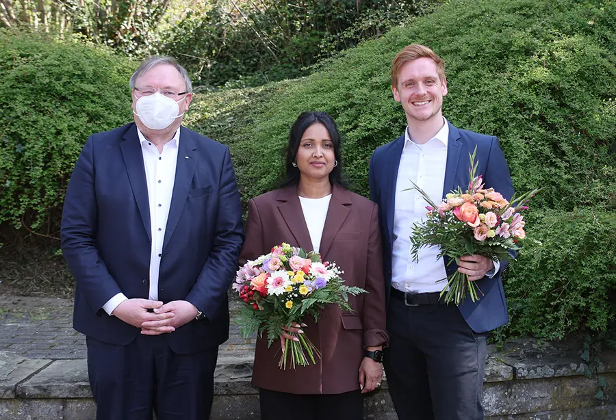 Roshani Thanapalasingham mit Blumenstrauß zwischen zwei Männern im Freien