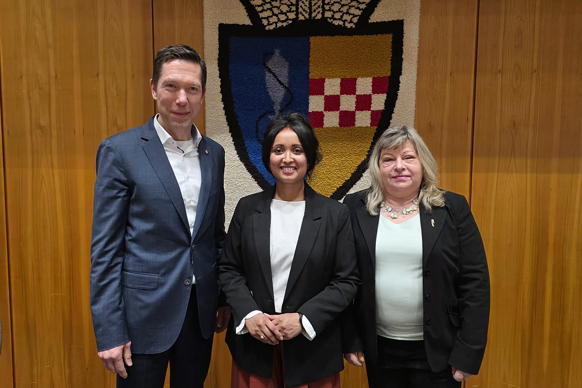 Roshani Thanapalasingham mit zwei weiteren Personen vor einem Wappen